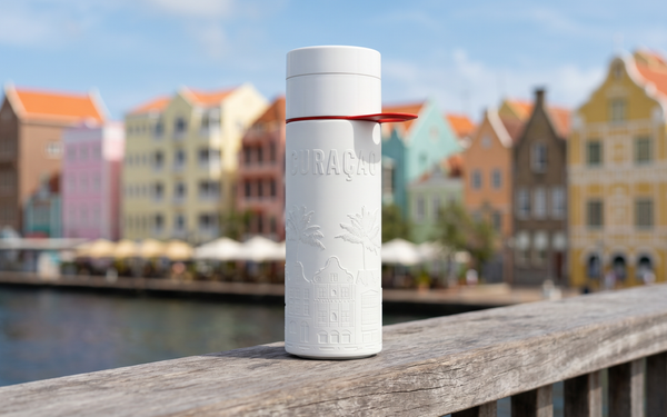 Herbruikbare Join the Pipe Curacao waterfles op een houten reling met de kleurrijke pastelhuizen van de Handelskade in Willemstad op de achtergrond.